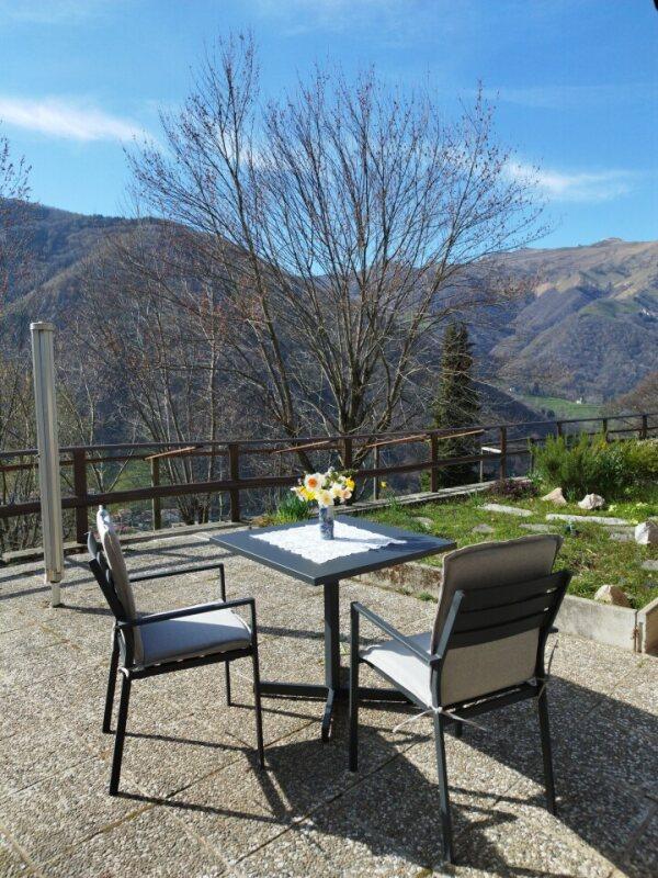 Ferienwohnung im Tessin - Terrasse mit Tisch und zwei Stühlen im Garten der Casa Natura mit Blick auf die Berge der Valle di Muggio
